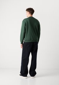Grønn genser laget av myk stoff, med rund hals og ribbestrikkede mansjetter, sammen med løse mørke jeans og hvite joggesko.