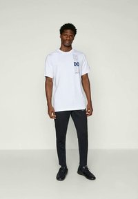 Biała koszulka graficzna z niebieskim napisem "JUST DO IT" i logo, połączona z czarnymi spodniami slim-fit. Model stoi w neutralnej przestrzeni.