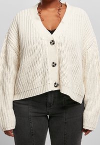 Krämfärgad, kraftigt stickad cardigan med V-hals, nedhasade axlar och svarta knappar. Har ribbade muddarna och nederkant, kort och boxig design.