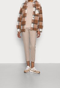 Chemise en flanelle à carreaux marron et blanc, hoodie beige, pantalon beige sur mesure et baskets marron avec accents blancs. Matériaux texturés et design superposé.