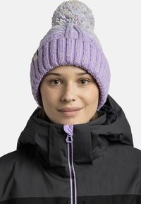 Knit beanie in light purple with a textured pattern and large pom-pom on top, worn with a black jacket featuring a zip closure.