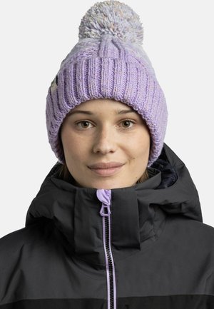 Gestrickte Mütze in Helllila mit einem strukturierten Muster und einem großen Pompon oben, getragen mit einer schwarzen Jacke mit Reißverschluss.