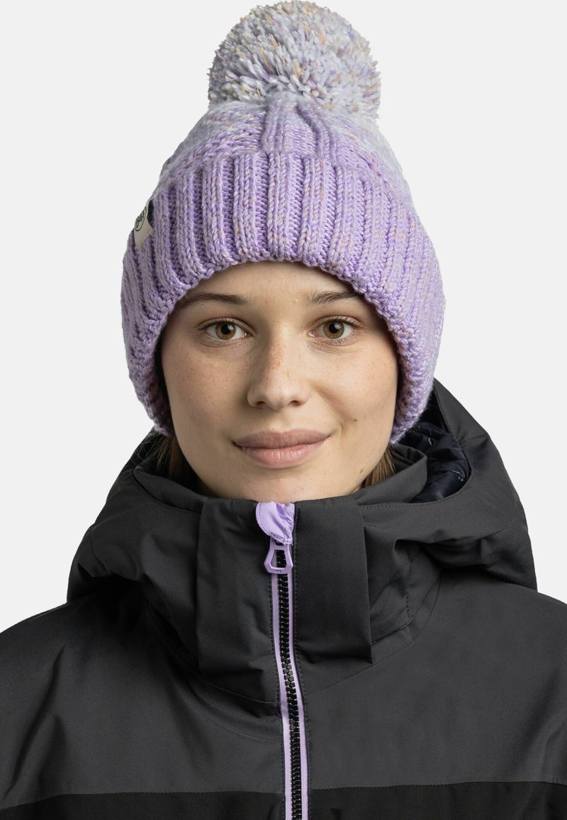 Knit beanie in light purple with a textured pattern and large pom-pom on top, worn with a black jacket featuring a zip closure.