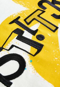 Gult stof med fede sorte broderede bogstaver og grafisk splattermønster. Inkluderer teksturerede detaljer og afrundede former.