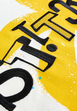 Gelbes Gewebe mit kräftigen schwarzen gestickten Buchstaben und grafischem Spritzmuster. Enthält strukturelle Details und abgerundete Formen.
