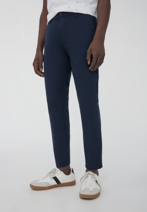 Pantaloni slim-fit blu navy con una texture liscia, dotati di passanti per cintura e chiusura a un bottone, abbinati a sneakers bianche.