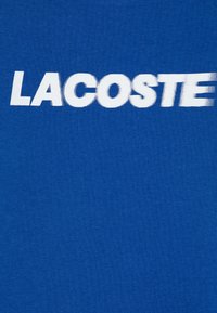 Tissu en coton bleu avec un grand texte blanc "LACOSTE" en police sans-serif en gras, avec une texture lisse et une couleur uniforme.