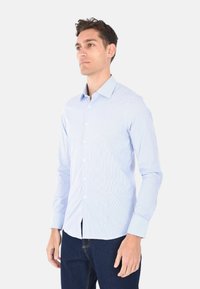 Camicia button-up a righe azzurro chiaro realizzata in cotone, con colletto classico, maniche lunghe e una vestibilità slim. Indossata con jeans scuri.