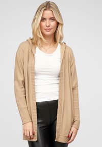 Beige cardigan met een capuchon, geribbelde mouwen en een open ontwerp, gedragen over een witte t-shirt en gecombineerd met zwarte broek. Zachte textuur.