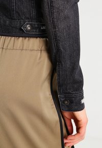 Giacca di jeans nera con una silhouette aderente, caratterizzata da bottoni metallici e dettagli usurati, abbinata a una gonna liscia di un beige chiaro.