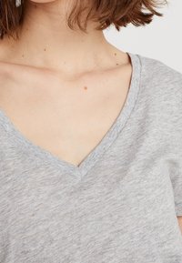 Primer plano del pecho superior y cuello de una persona con piel clara y pecas, vistiendo una camiseta de cuello en V de color gris claro.