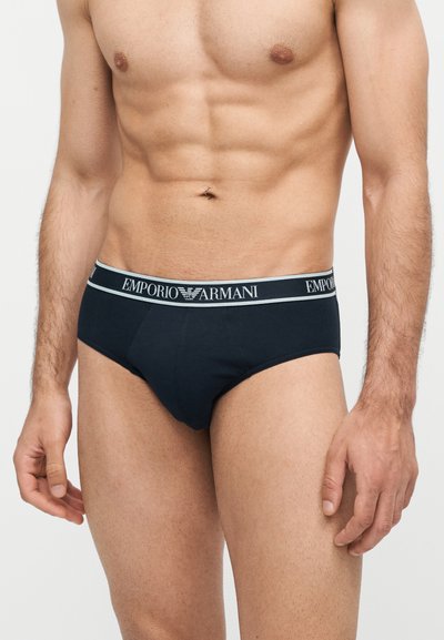 Μυώδης ανδρικός κορμός που φοράει μαύρα εσώρουχα Emporio Armani με λογότυπο στη μέση, στέκεται μπροστά σε απλό φωτεινό φόντο.