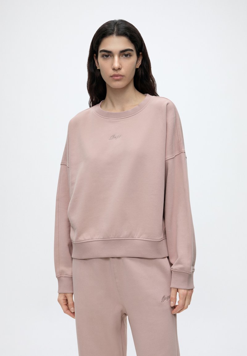 Femme aux longs cheveux foncés portant un sweat-shirt ample mauve clair et un pantalon de jogging assorti sur un fond blanc uni.