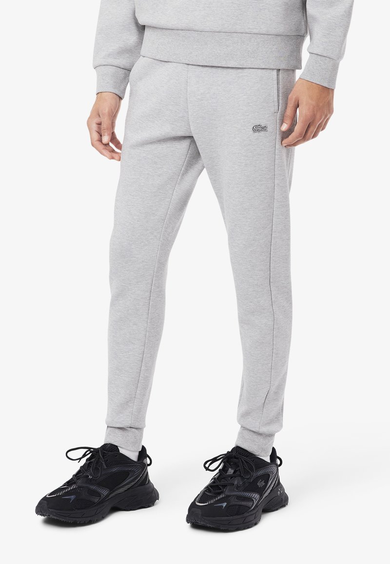 Pantalones de chándal grises hechos de una tela suave, con piernas ajustadas, detalles en las costuras laterales y un pequeño logo en la cadera. Combinados con zapatillas de deporte negras.