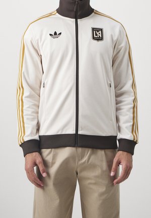 Chaqueta atlética blanca de poliéster con tres rayas doradas, cuello y puños marrones, bolsillos con cremallera y logo del Los Angeles FC en el pecho.