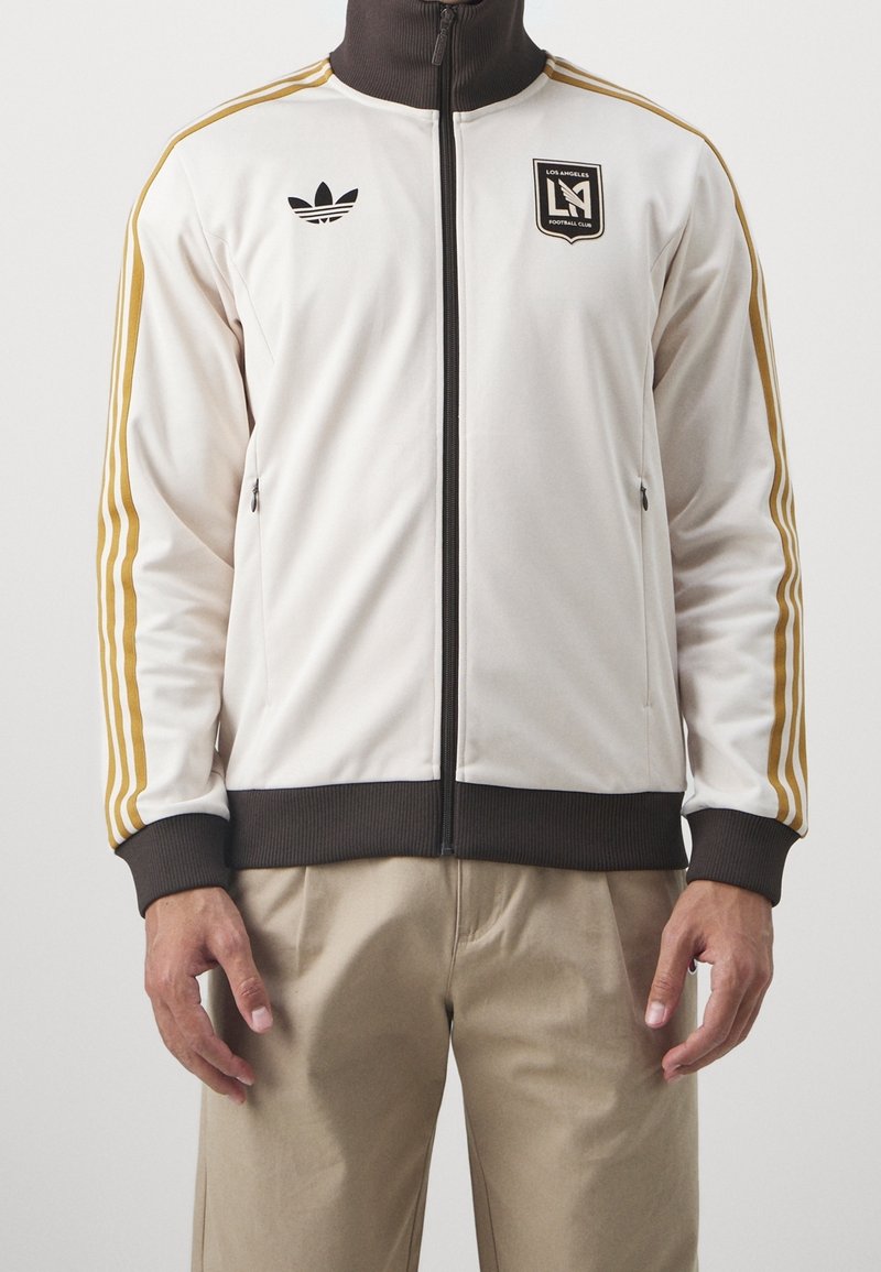 Bílá polyesterová sportovní bunda se zlatými trojitými pruhy, hnědým límcem a manžetami, zipovými kapsami a logem Los Angeles FC na hrudi.