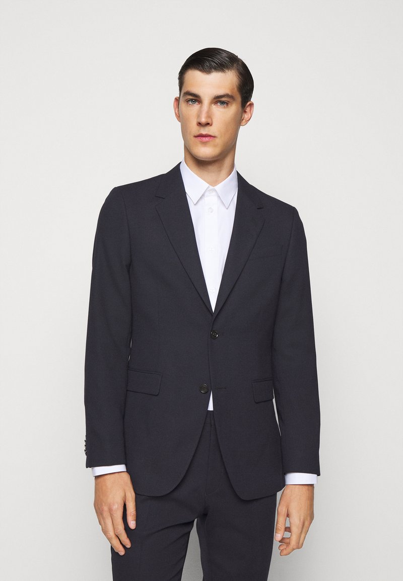 Blazer azul marinho feito de tecido, com fecho de dois botões, lapelas em bico, bolsos laterais e silhueta estruturada, combinado com uma camisa branca.