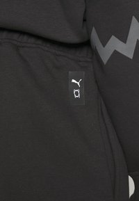 Čierna mikina s kapucňou s nášivkou s bielym logom Puma a grafikou úst. Tkanina je hladká s jemnou textúrou, čo zdôrazňuje dizajn.