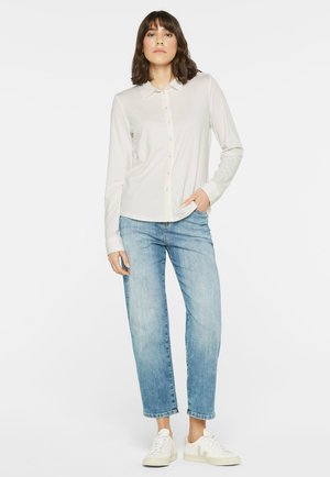 Frau steht mit einer Hand in der Tasche, trägt ein weißes Hemd mit Knöpfen, hellblaue Jeans und weiße Turnschuhe vor einem schlichten weißen Hintergrund.