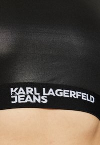 Musta joustava rintaliivi, jossa on sileä pinta. Alareunassa on valkoinen "KARL LAGERFELD JEANS" -brändi. Minimalistinen muotoilu, ei lisäkuvioita.