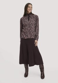 Langärmliges Hemd mit einem pinken und schwarzen Tiermuster, kombiniert mit einem dunkelbraunen Rock. Das Modell trägt braune Stiefel bis zum Knie.