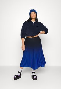 Ναυτικό cropped φλις πουλόβερ με κλείσιμο φερμουάρ, συνδυασμένο με πλισέ μπλε gradient φούστα και μαύρες πλατφόρμες. Το μοντέλο φορά επίσης λευκές κάλτσες.