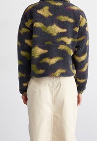 Polarowa bluza w ciemnoszarym kolorze z kamuflażem w odcieniach zieleni i żółci. Posiada luźny krój i zaokrąglone szwy na ramionach.