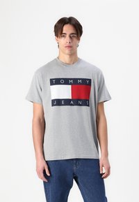 Hallikas puuvillane T-särk, millel on värviline plokkdisain, millel on tumesinine, valge ja punane logo print "TOMMY JEANS." Lühikesed varrukad ja ümmargune kaelus.