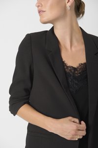 Blazer nero dal taglio sartoriale, con rever strutturati e maniche arrotolate, indossato sopra un top in pizzo nero. Lucentezza sottile, design elegante.