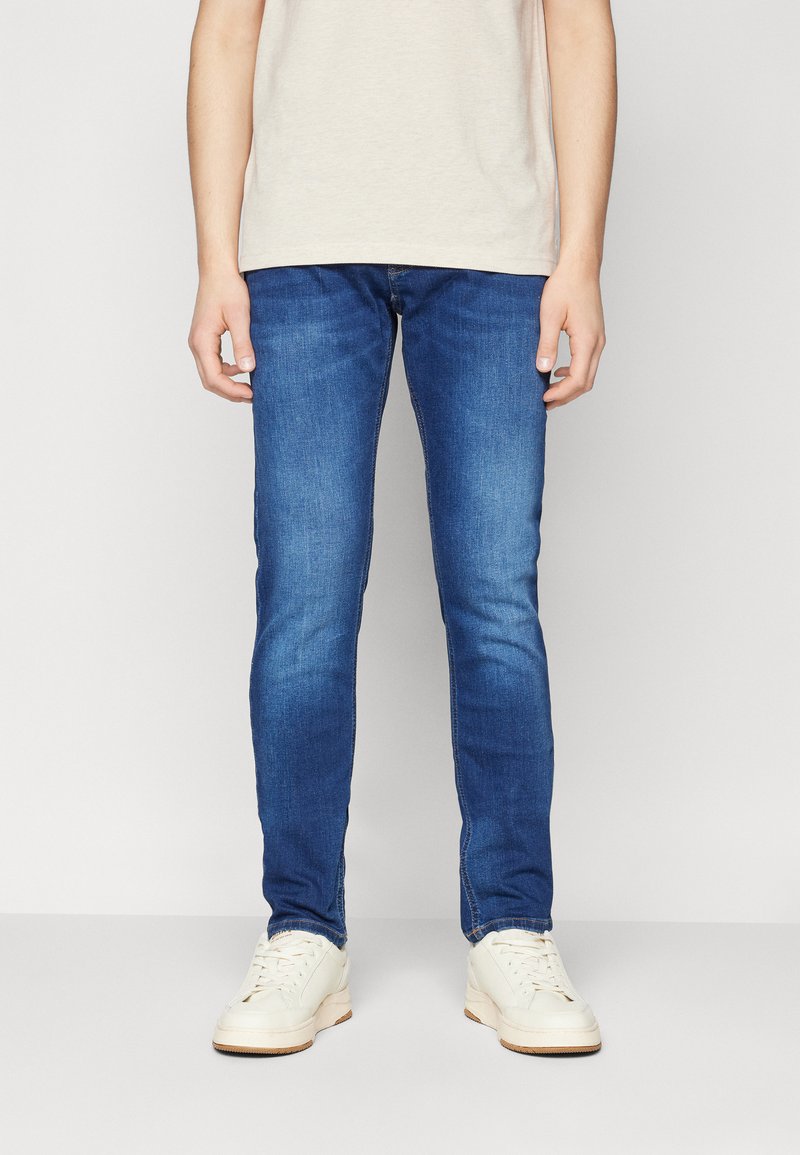 Slim fit kék farmernadrág, fehér sportcipővel és világos bézs, rövid ujjas pólóval, egy sima fehér háttér előtt állva.