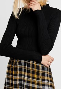 Top lungo a maniche nere a costine con collo alto. Abbinato a una gonna a quadri gialla, nera e bianca, con un design plissettato.