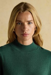 Roheline kudumiga särk, millel on kõrge ribatud krae ja tekstuuriga kangas, mida kannab pikkade juustega mudel neutraalses taustas.