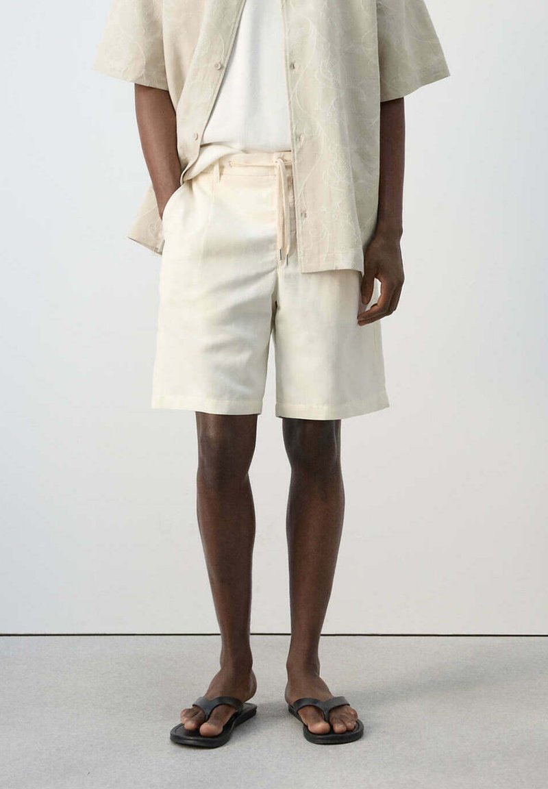 Modèle masculin portant un short beige clair avec cordon, une chemise à manches courtes assortie avec boutons, un t-shirt blanc et des sandales noires.