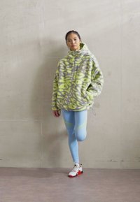 Casaco polar em verde limão e cinza com padrão abstrato; leggings azul claras com uma faixa amarela; sapatilhas brancas desportivas com detalhes vermelhos.