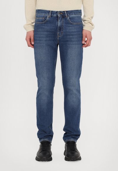 Jean en denim bleu avec une coupe droite, cinq poches et une finition délavée. Porté avec des baskets noires et un haut à manches longues de couleur claire.