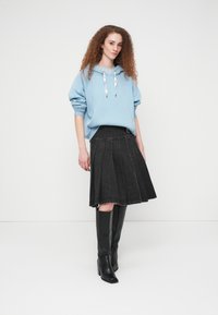 Νεαρή γυναίκα με σγουρά μαλλιά, φορόντας ένα ανοιχτό γαλάζιο φούτερ με κουκούλα, μαύρη πλισέ φούστα και μαύρες μπότες μέχρι το γόνατο, μπροστά σε απλο φόντο λευκού χρώματος.