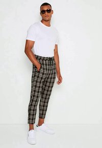Homem vestindo calças quadriculadas em preto e branco, t-shirt branca, ténis brancos e óculos de sol escuros, de pé com uma mão no bolso.