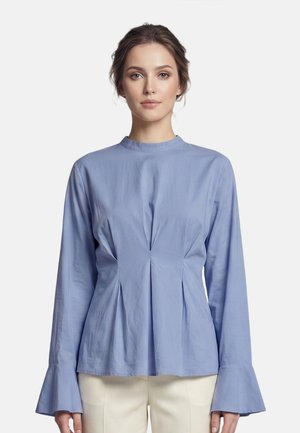 Vrouw draagt een lichtblauwe blouse met lange mouwen, uitlopende manchetten en plooien aan de voorkant, gecombineerd met crèmekleurige broek.