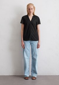Zwarte blouse met korte mouwen en een V-hals, gecombineerd met lichtblauwe straight-leg jeans. Model staat tegen een lichtgeklede, gestructureerde muur.
