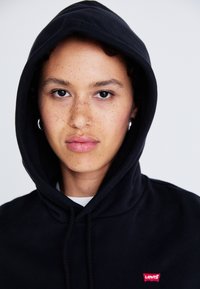 Čierna mikina s kapucňou vyrobená z mäkkej látky, so predným klokankovým vreckom a malým červeným logom Levi's na hrudi.