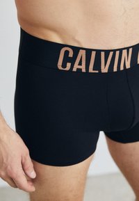 Schwarze Boxershorts aus dehnbarem Material; ausgestattet mit einem breiten, beigen Bund, auf dem "CALVIN" in fetter Schrift steht. Glattes, enganliegendes Design.