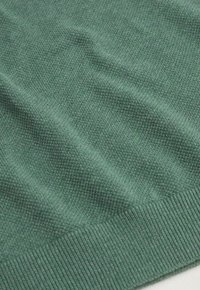 Gros plan sur un pull en tricot vert, présentant un tissu texturé avec un ourlet côtelé, affichant un motif de tissage subtil et régulier.