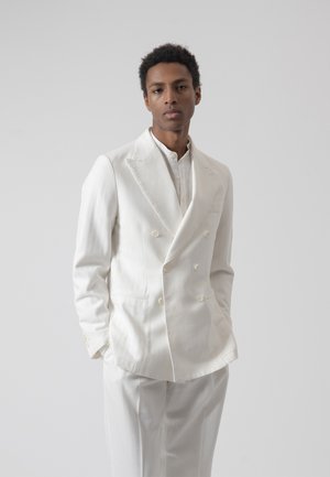 Hombre con traje blanco cruzado y pantalones a juego, de pie con las manos en los bolsillos sobre un fondo gris liso.