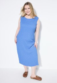 Vestido azul sin mangas, de largo hasta la rodilla, con una tela texturizada que presenta un distintivo patrón arrugado. Combinado con zapatos marrones tipo slip-on.
