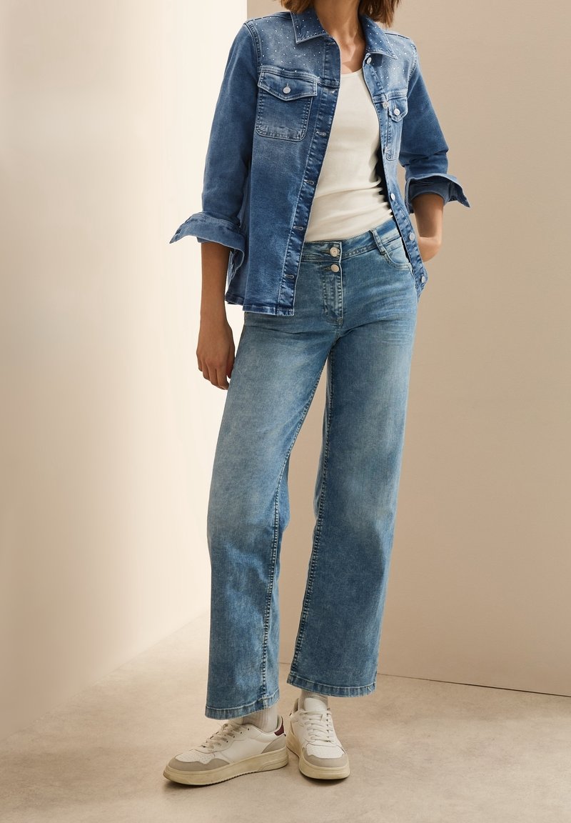 Femme portant un jean taille haute bleu clair, des baskets blanches, un haut blanc et une veste en jean bleu foncé aux manches retroussées sur fond neutre.