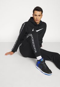 Felpa nera con logo Nike bianco, pantaloni jogger neri abbinati con strisce laterali bianche e sneakers alta blu/nera con suole bianche.