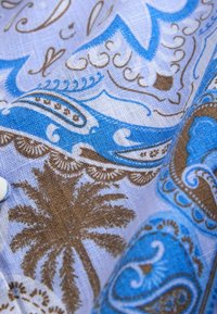 Tissu avec des motifs cachemire bleus et bruns et des palmiers sur un fond clair, montrant une texture plissée et des fils visibles.