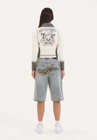 Suéter de punto blanco y gris con gráfico de calavera y letras en la parte posterior, combinado con pantalones cortos de mezclilla claros que presentan un bordado colorido de dragón.