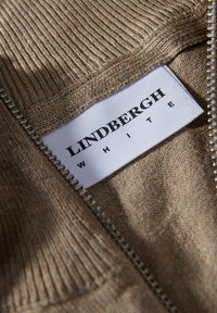 Beige stickad tröja med ribbat mönster, med en silverdragkedja och en vit etikett som visar "Lindbergh White" i fet svart font.