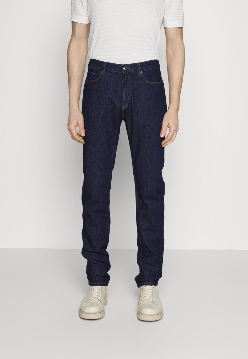 Mörkblå denimjeans med slim fit, med traditionella fickor och sömmar. Bärs med en vit, texturerad t-shirt med kort ärm och krämfärgade skor.